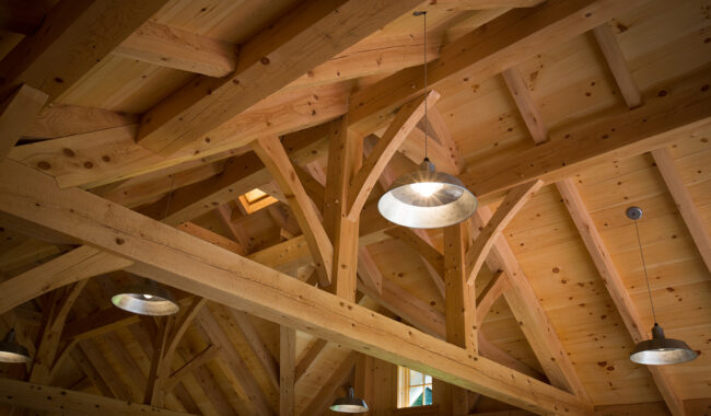 Exposed wooden beams form the ceiling structure of a building, with several metal pendant lights hanging down and sunlight shining through small windows and skylights.