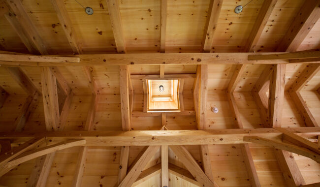A view looking up at a wooden vaulted ceiling with exposed beams and a small square skylight in the center, allowing natural light to enter the room.