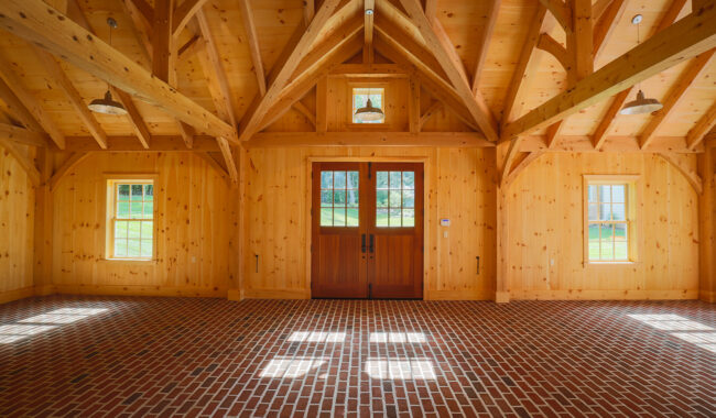 Spacious room with wood beams, paneled walls, a brick floor, and double glass doors. Sunlight streams through windows, casting patterns on the floor. The room has a rustic, warm ambiance.
