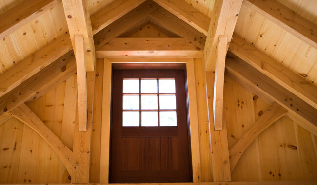 A close-up view of a wooden loft or attic with exposed beams and a small windowed door centered on the wall, letting in natural light. The interior is made entirely of light-colored wood.