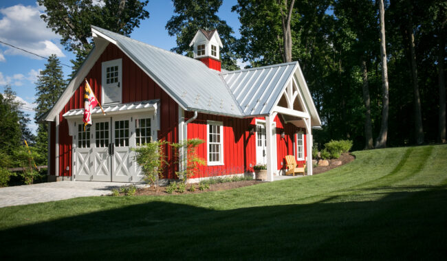 A red barn-style building with white trim, metal roof, and a small cupola, surrounded by lush green grass and trees. An American flag and an Adirondack chair are near the front entrance.