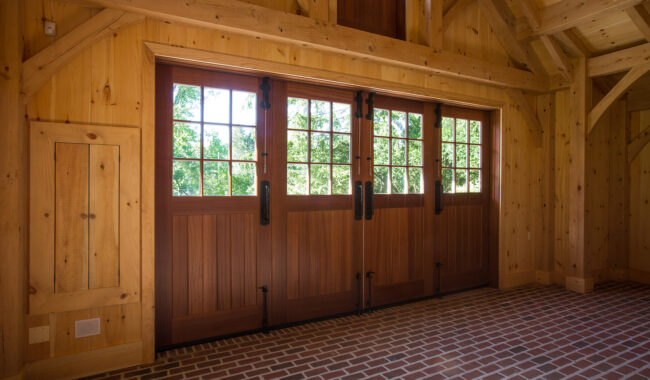 A wooden garage with large wooden doors featuring glass window panes, exposed timber beams, and a red brick floor. Sunlight streams through the windows, highlighting the natural wood interior.