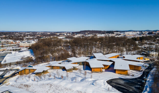 Aerial view of a modern building with angular roofs covered in snow, surrounded by trees and a snowy landscape, with a town visible in the background under a clear blue sky.
