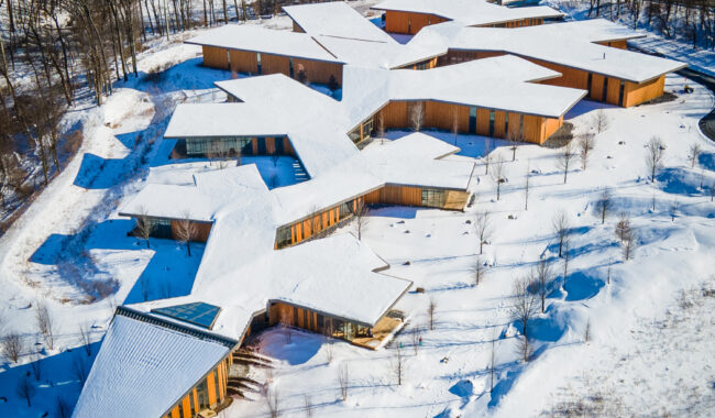 Aerial view of a large, modern building with an angular, star-shaped design, covered in snow and surrounded by a snowy landscape with scattered trees and nearby houses.