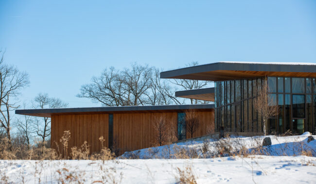 A modern building with large glass windows and wooden walls sits on a snowy landscape under a clear blue sky, with leafless trees in the background.