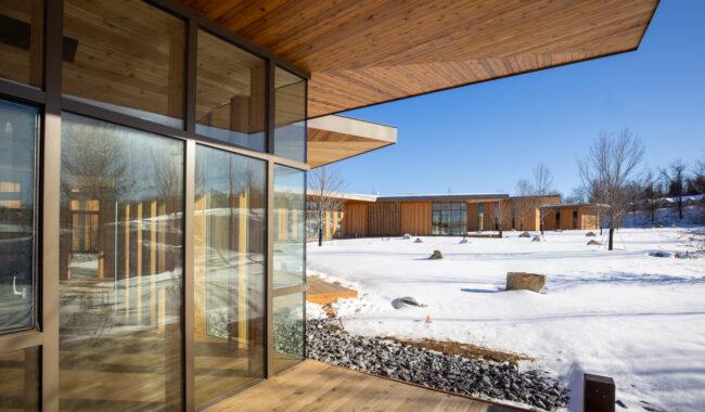 A modern building with large glass windows and wooden overhangs overlooks a snowy landscape with scattered rocks and bare trees under a clear blue sky.