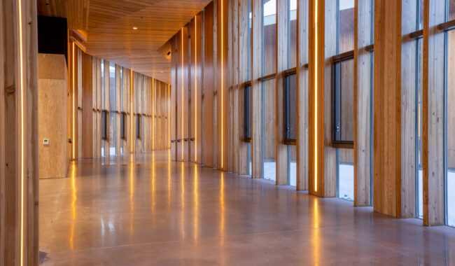 A modern corridor with tall wooden beams, floor-to-ceiling windows, and polished concrete floors. Warm lighting highlights the wood, creating a bright, inviting atmosphere.