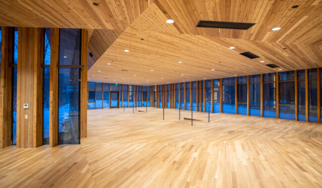 Spacious room with floor-to-ceiling windows and a modern wood-paneled ceiling and floor. The area is empty, and natural light streams in, highlighting the warm wood tones and minimalist architectural design.