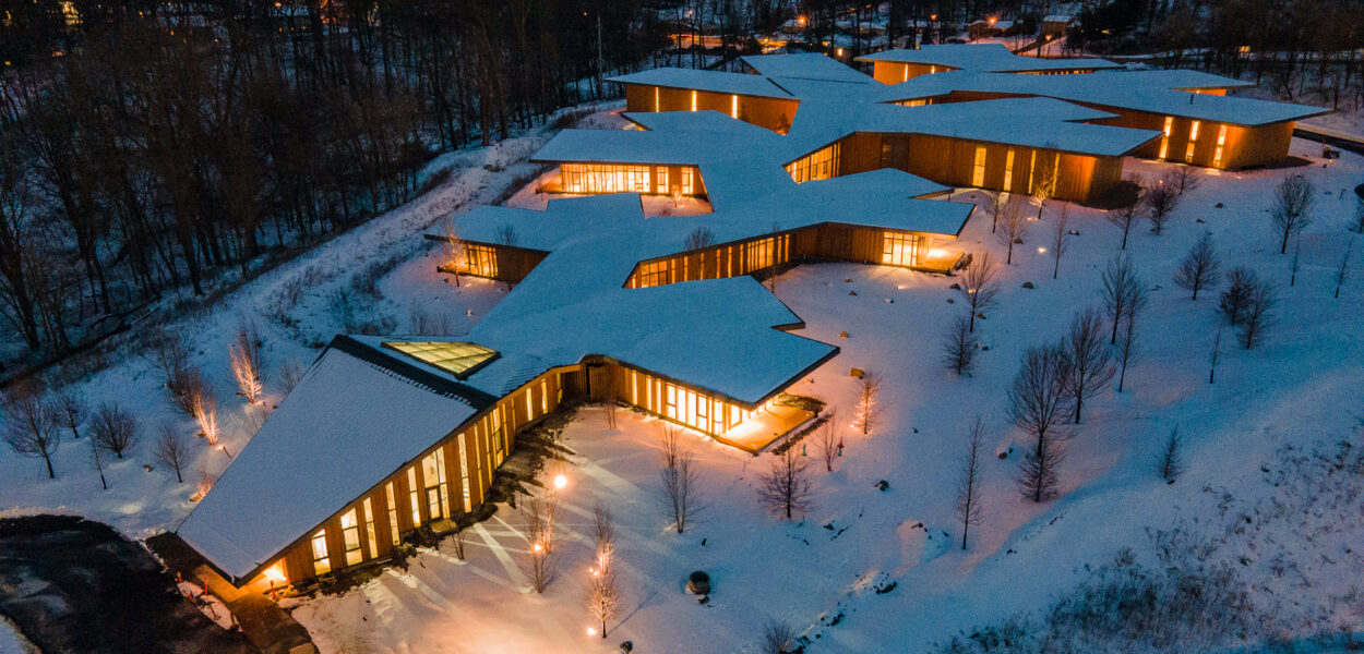 Aerial view of a modern building with angular, branching design, illuminated from within at dusk, surrounded by snow-covered ground and bare trees.