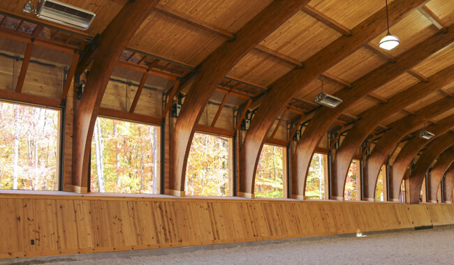 Interior of a wooden riding arena with large arched beams, open windows showing autumn trees outside, and sunlight streaming in. The ground inside is covered with light-colored sand or gravel.
