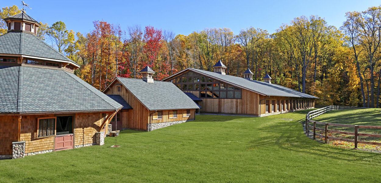 A large wooden barn complex with green lawns, surrounded by a wooden fence and colorful autumn trees under a clear blue sky.