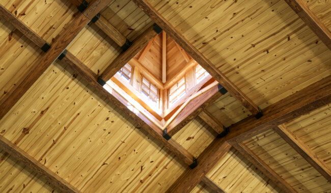 Wooden ceiling with intersecting beams and a square opening in the center, revealing natural light coming through windows above. The wood has a warm, natural finish, creating a cozy architectural detail.