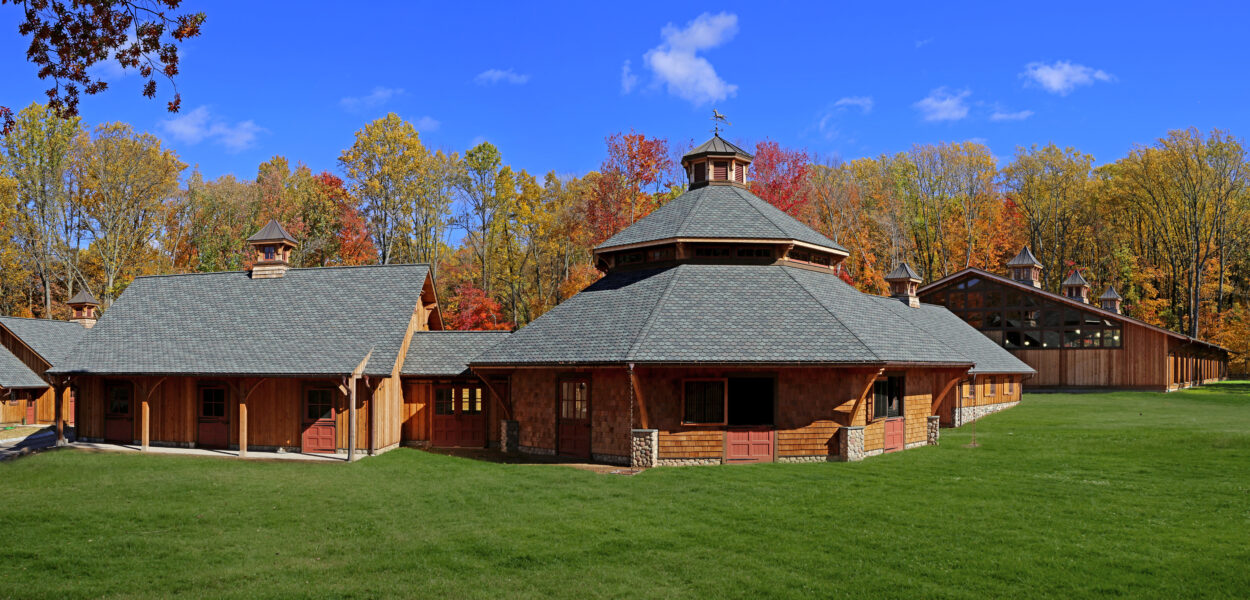 A large wooden stable complex with a round central building and pitched roofs stands on a green lawn, surrounded by autumn trees under a clear blue sky.