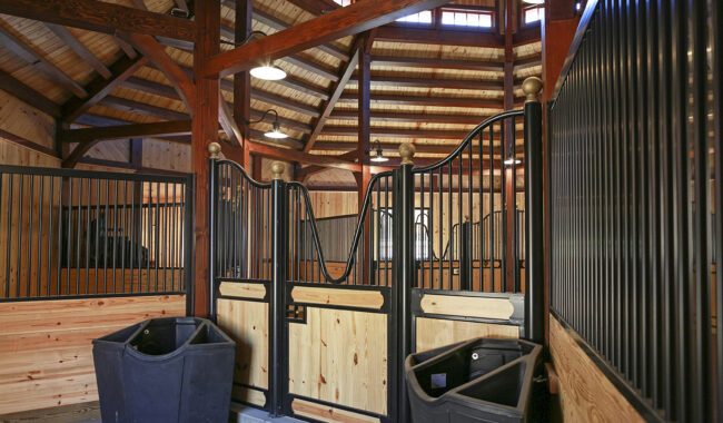 Interior of a well-lit, wooden horse stable with metal railings, wood paneling, and two horse stalls, each with a black feed bin in front. The ceiling features exposed beams and hanging lights.