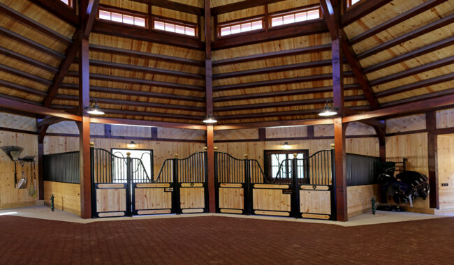 A spacious, octagonal horse stable with high wooden ceilings, exposed beams, and brick flooring. Four horse stalls with black metal gates line the back wall. Tack and cleaning supplies are organized on the right side.