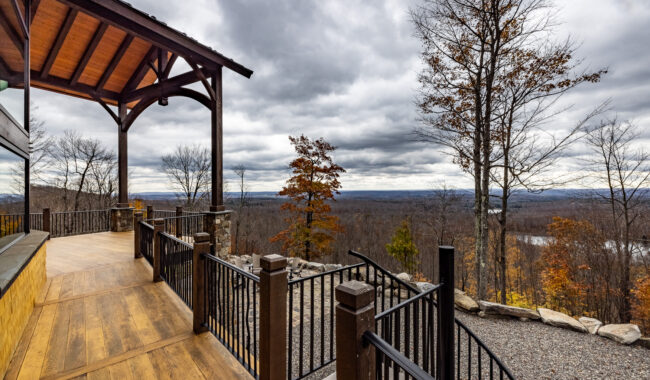 A covered wooden deck with black railings overlooks a scenic, tree-filled landscape and distant river under a cloudy sky, creating a tranquil outdoor atmosphere.
