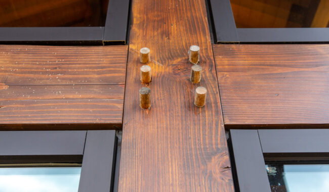 Close-up of a wooden beam joint with six round wooden dowels securing the intersecting beams. The beams have a rich brown finish, and black-framed glass windows are visible in the background.