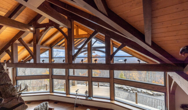 A spacious room with a high wooden ceiling and large triangular windows offers a scenic view of mountains and trees. There is a telescope by the window, cozy seating, and a wooden deck outside.