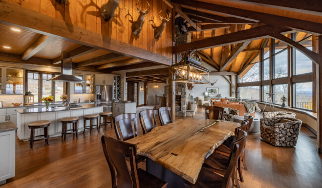 A spacious rustic kitchen and living area with wooden beams, a live-edge dining table, mounted deer heads, large windows overlooking scenery, and modern furnishings. Warm lighting and earth tones create a cozy atmosphere.