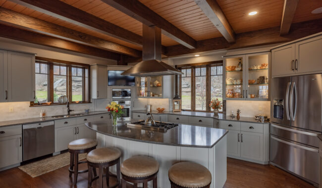 Modern kitchen with large island, four stools, stainless steel appliances, gray cabinets, open shelves with dishware, and large windows letting in natural light. Exposed wood beams on the ceiling add warmth to the space.