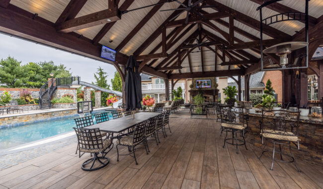 Covered outdoor patio with wooden beams, ceiling fans, and bar seating overlooking a swimming pool. Tables and chairs are arranged under the roof, with greenery and a TV visible in the background.
