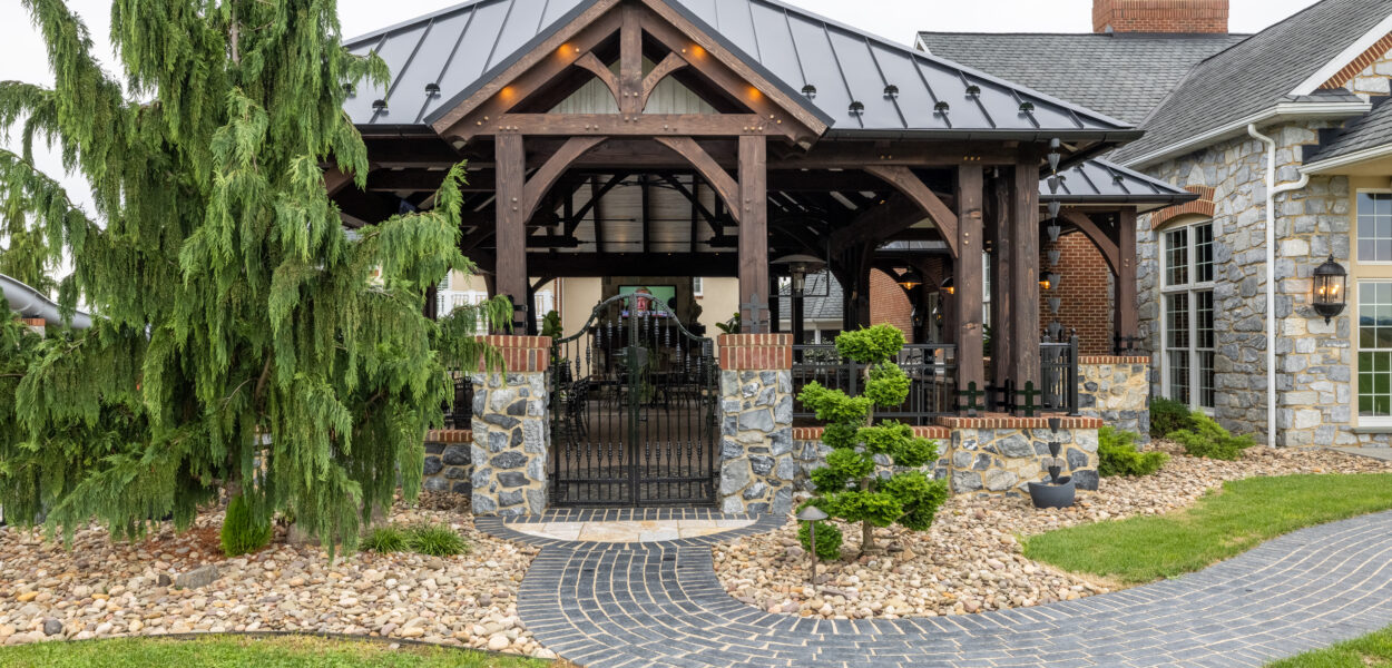 A covered outdoor patio with exposed wooden beams, stone columns, and a black metal roof, surrounded by landscaped rocks, a curved stone walkway, and green shrubs next to a brick and stone house.
