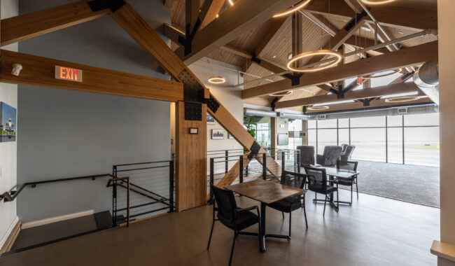 A modern, open interior with exposed wooden beams, circular ceiling lights, black metal railings, and glass walls. Several tables and chairs are arranged near large windows facing an outdoor area.