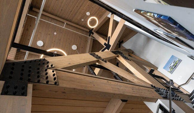View looking up at a modern wooden ceiling with exposed beams and black metal brackets, circular light fixtures, and white walls with a framed artwork on the right side.