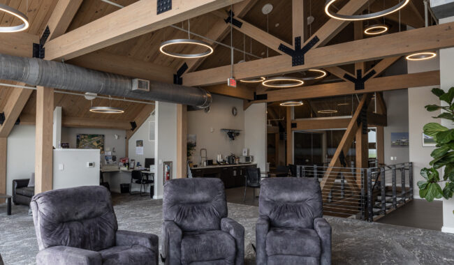 Modern lounge area with gray recliner chairs, a round white table, exposed wooden beams on a vaulted ceiling, and circular pendant lights hanging above. An office kitchen and staircase are visible in the background.