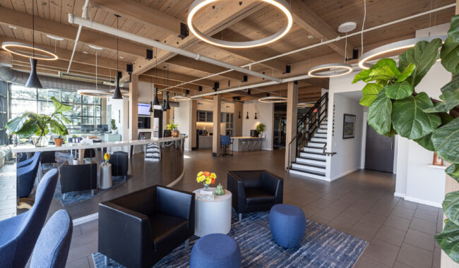 Modern office lobby with wooden ceiling, circular pendant lights, black armchairs, blue ottomans, a blue rug, potted plants, and a staircase. Large windows provide natural light and an open, welcoming atmosphere.