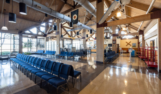 A spacious, bright room with high wooden ceilings, blue chairs arranged in rows, large windows, and aviation-themed decor including model planes and banners. The area appears to be a waiting or event space.