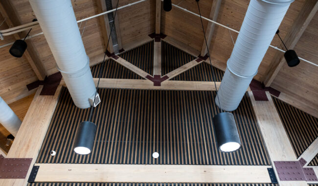 View of a modern wooden ceiling with exposed beams, large cylindrical air ducts, hanging black pendant lights, and striped paneling creating a geometric pattern.