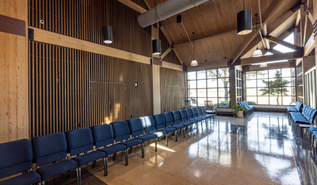 A spacious room with high wooden ceilings, large windows letting in sunlight, and rows of blue cushioned chairs arranged along the walls. The space appears clean, modern, and inviting.