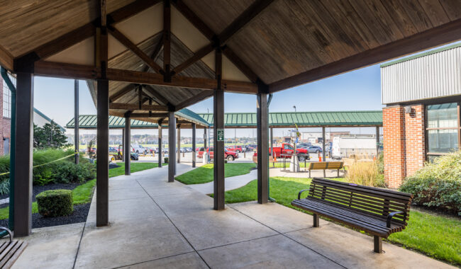 A covered outdoor walkway with wooden beams and a bench leads to a parking lot with cars. Green lawns, landscaped bushes, and adjacent buildings are visible on a sunny day.