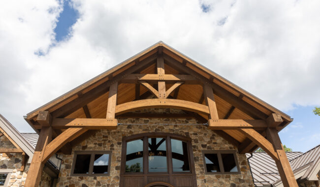 The image shows the front entrance of a stone house with large wooden beams forming a peaked roof. There are several windows and the sky above is partly cloudy.