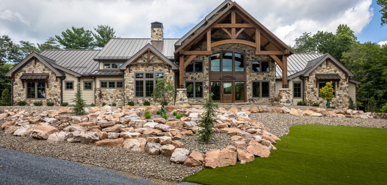 A large, rustic house with stone and wood exterior, tall windows, a peaked roof, and timber beams. The landscaped front yard features rocks, mulch, small trees, and a patch of artificial grass.