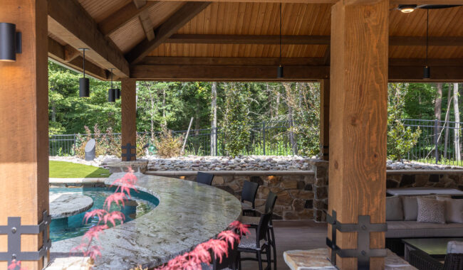Covered outdoor patio with wooden beams, a curved stone bar with barstools, and a view of a pool and green trees in the background. The area is shaded, with lounge seating visible to the right.