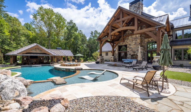 A luxurious backyard features a curved swimming pool with a hot tub, stone patio, lounge chairs, and a large rustic house with timber beams and a covered outdoor seating area surrounded by lush trees under a partly cloudy sky.