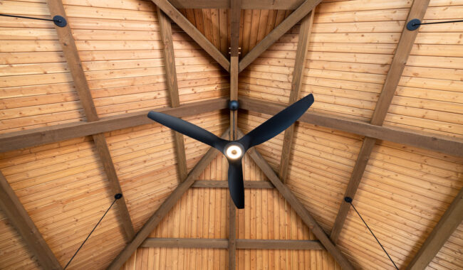 A modern black ceiling fan with three blades is mounted on a high, wooden, pitched ceiling with exposed beams and warm-toned wood panels.