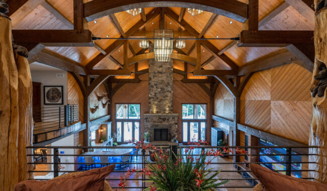 Spacious lodge-style living room with high vaulted wooden ceilings, exposed beams, large stone fireplace, tall windows, modern lighting, and a plant arrangement in the foreground.