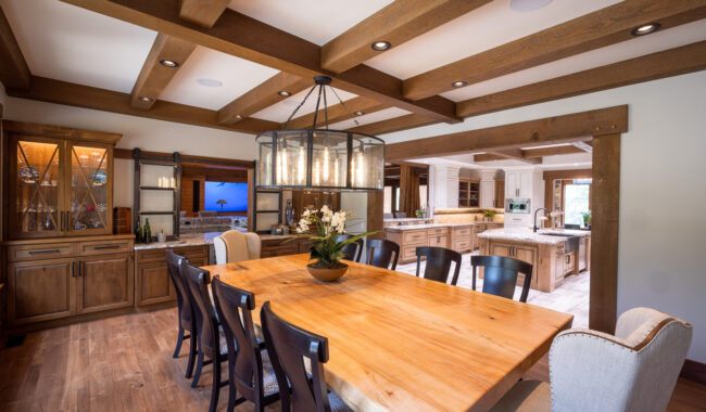 Spacious dining room with a large wooden table, black and beige chairs, modern chandelier, wooden ceiling beams, glass cabinets, and an open view into a bright kitchen.