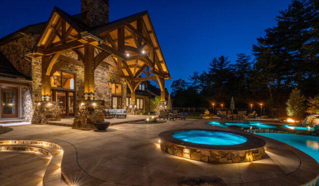Luxurious stone house with large wooden beams, illuminated patio, pool, and hot tub at night. Outdoor seating and landscaped greenery surround the brightly lit water features.