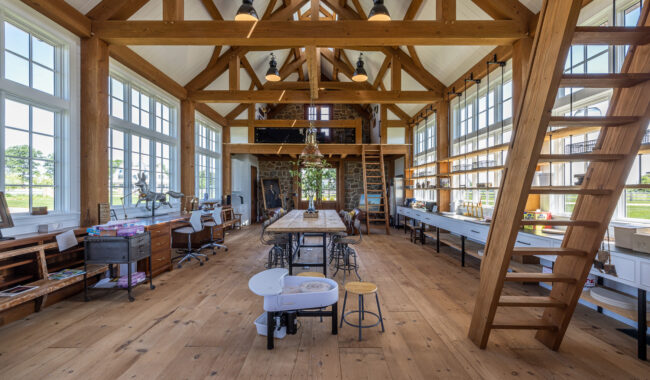 Spacious, sunlit art studio with exposed wooden beams, large windows, long worktables, shelves with art supplies, and a wooden ladder leading to a loft. Light wood floors enhance the bright, airy atmosphere.