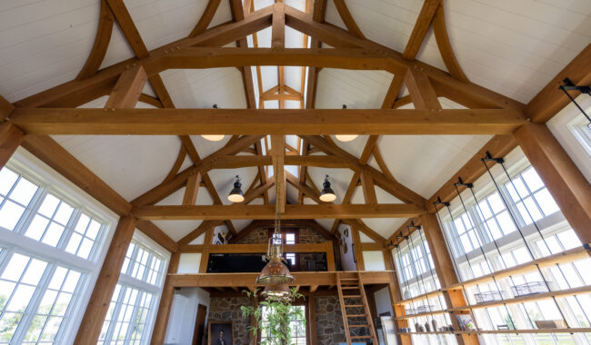 Interior view of a spacious room with exposed wooden beams, large windows letting in natural light, a loft area with a ladder, and shelves along one wall. The ceiling is vaulted and white.