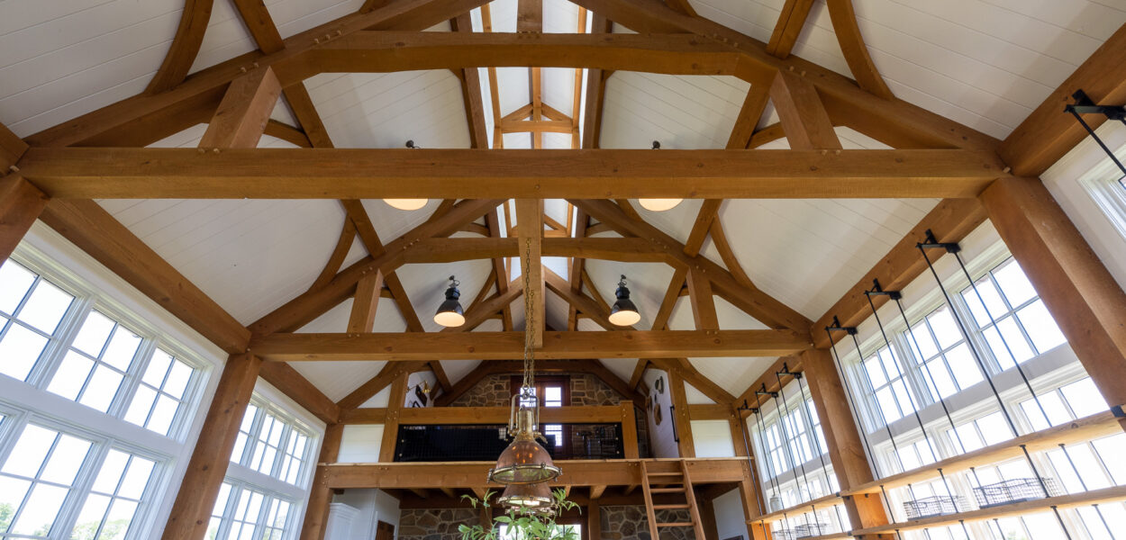 Interior of a spacious room with exposed wooden beams, vaulted ceiling, large windows, hanging lights, a loft area with a ladder, and a kitchen with open shelving on the right. Natural light fills the space.