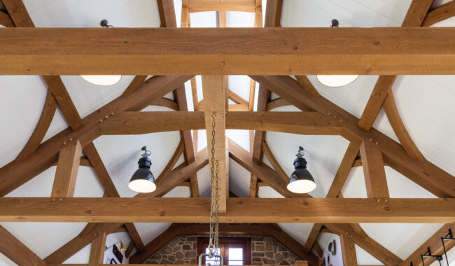 Exposed wooden beams and trusses support a vaulted white ceiling with black light fixtures hanging down, creating a rustic, open atmosphere in the room.