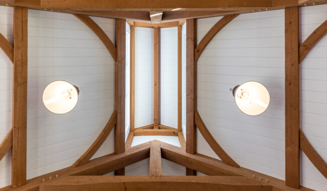 View of a vaulted ceiling with exposed wooden beams and white shiplap panels, featuring two round light fixtures evenly spaced along the ceiling.