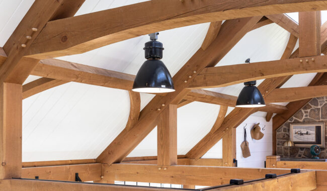 Exposed wooden beams and black pendant lights hang from a vaulted ceiling with white paneling; a stone wall with a framed picture and hanging hats is visible in the background.
