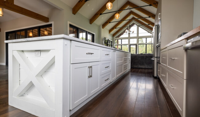 Modern kitchen with a large white island, wooden floor, exposed wooden ceiling beams, pendant lights, and large windows letting in natural light.