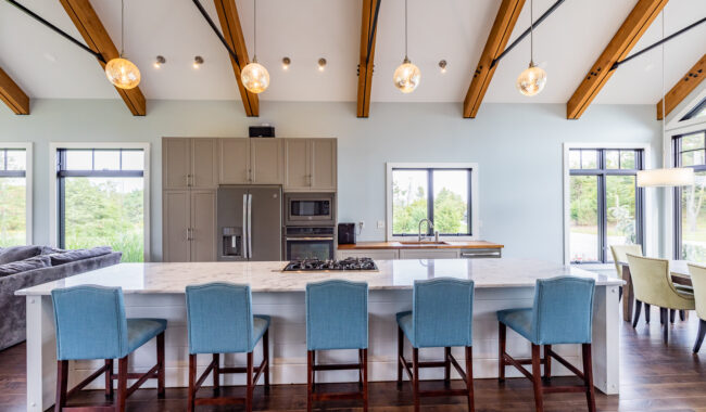Modern kitchen with exposed wooden ceiling beams, pendant lights, a large island with a marble countertop, five blue chairs, stainless steel appliances, and windows letting in natural light.